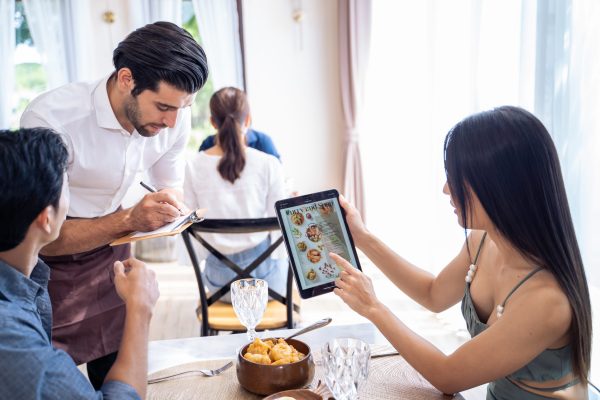 Caucasian waiter receiving order serving from customer in restaurant. Attractive server service man working, taking note, writing order from consumer with pen at table in dining room with happiness.