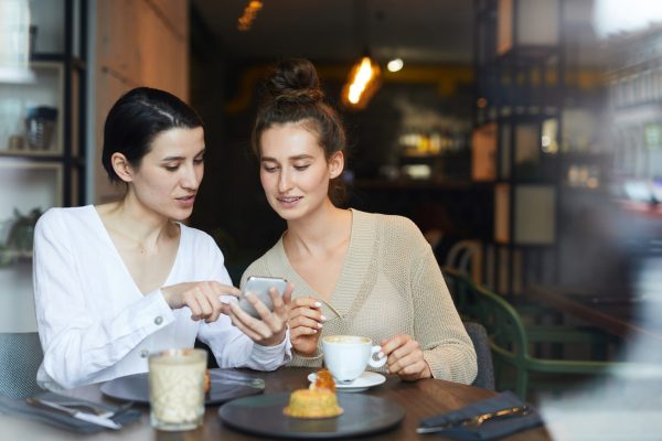 Two friendly casual girls relaxing in cafe, having coffee with dessert and discussing curious looking in smartphone