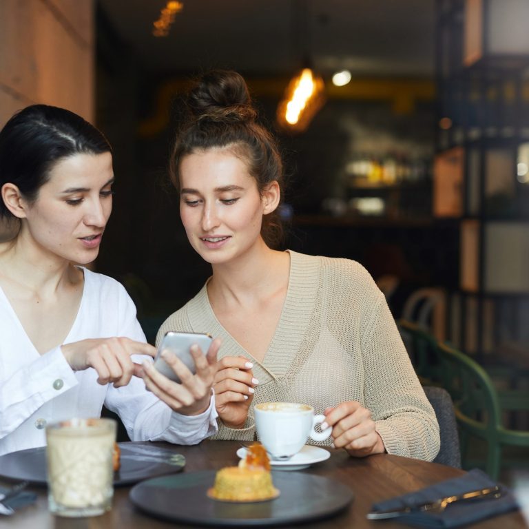 Two friendly casual girls relaxing in cafe, having coffee with dessert and discussing curious looking in smartphone